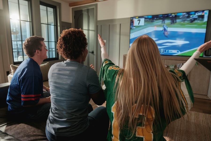 Friends in jerseys watch a football game together on a mounted TV in a modern living room, one raising her arm to celebrate.